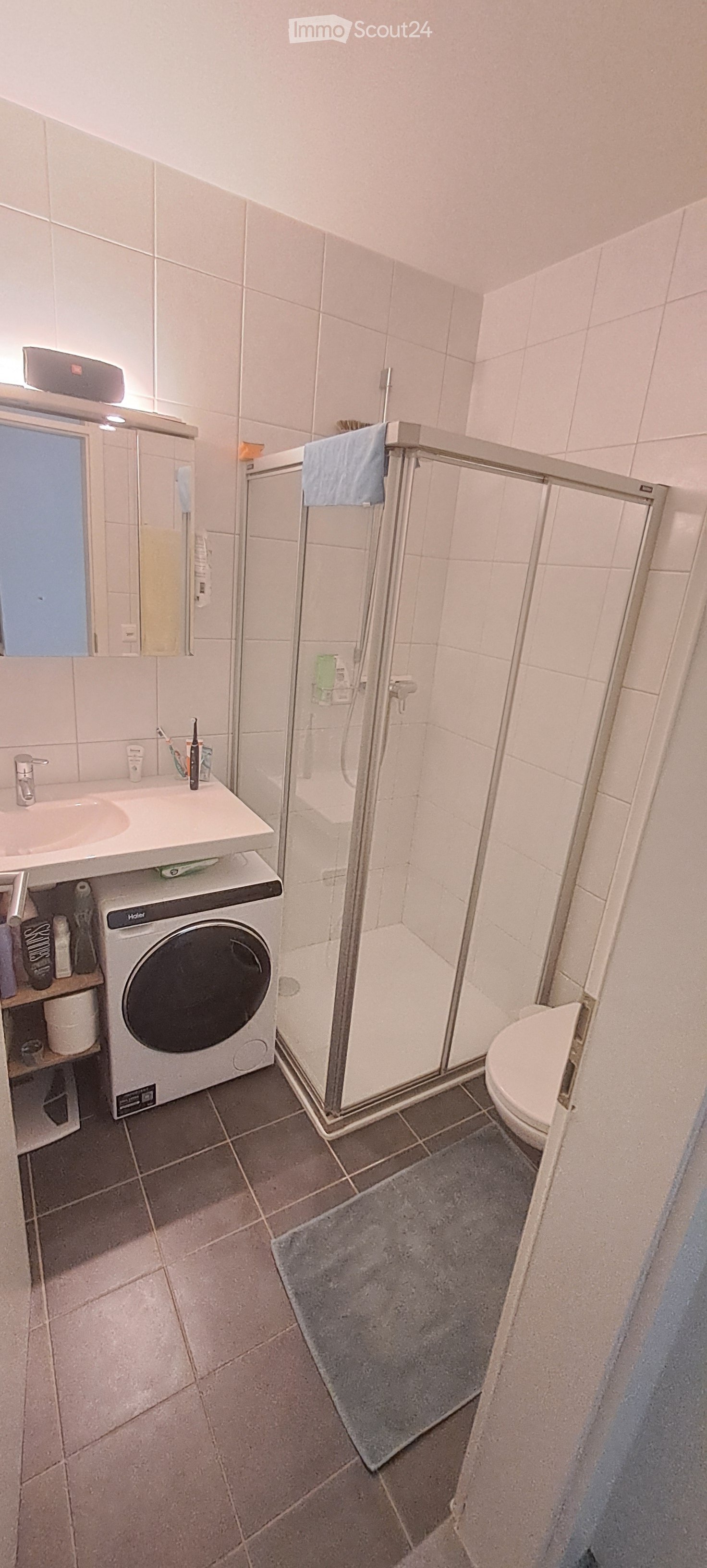 Modern bathroom with white tiles, a sink with a mirror, a shower with a glass door, a toilet, and a washing machine. A towel hangs on the shower door, and a bathmat is placed on the floor.