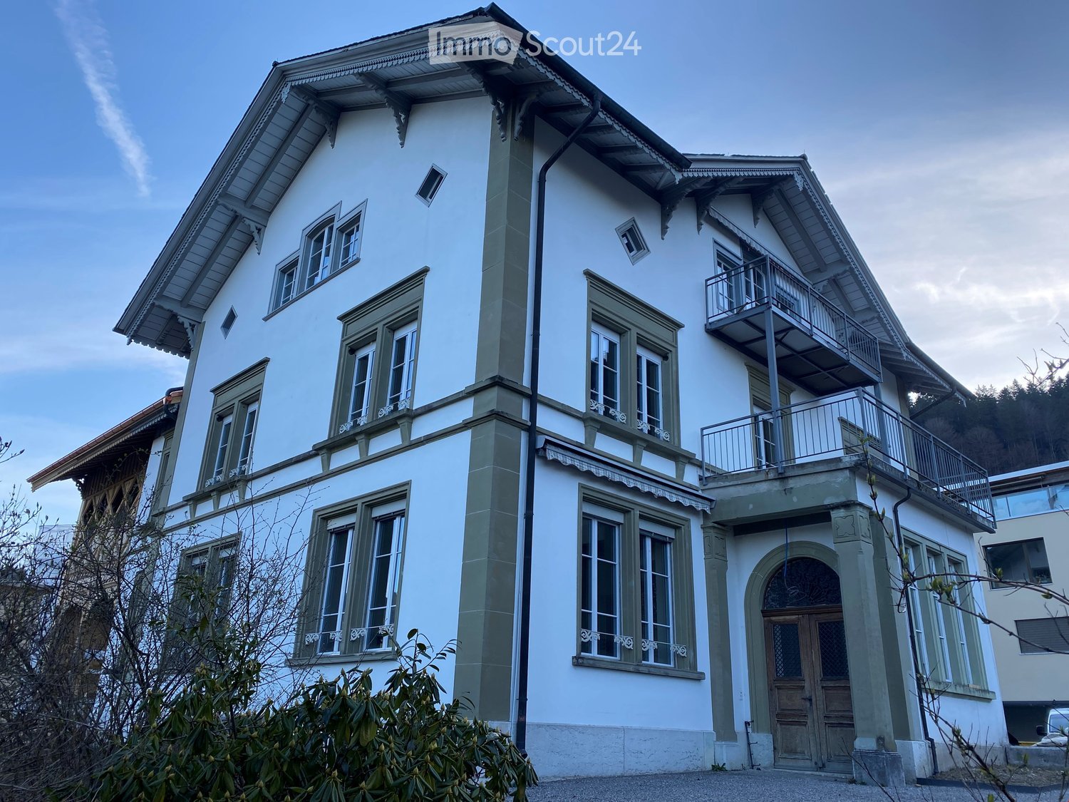 A two-story white house with green-framed windows, a balcony on the upper floor, and decorative architectural details.