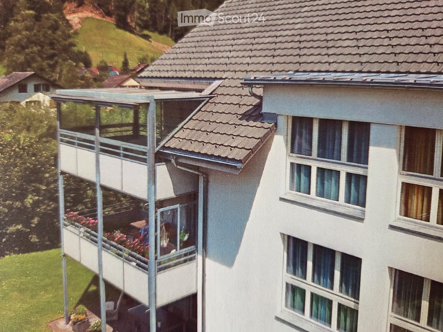 A multi-story residential building with a tiled roof, large windows, and a balcony area with railings and potted plants.