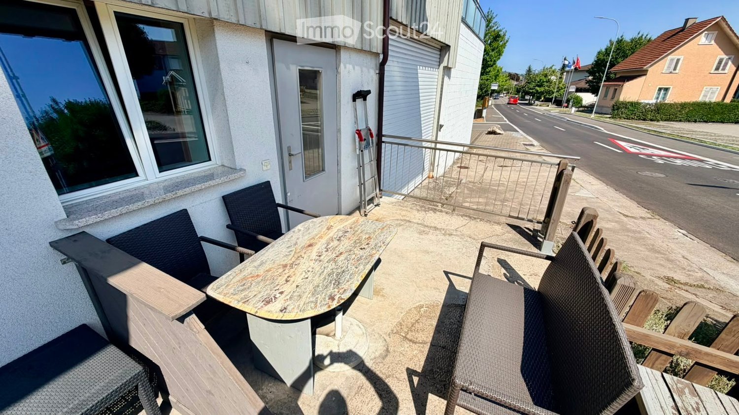 Balcony with a table and chairs, overlooking a street with houses and trees.