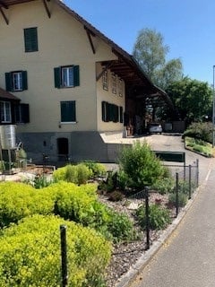 A two-story building with a beige facade and green shutters, surrounded by a well-maintained garden with neatly trimmed bushes and a gravel pathway.