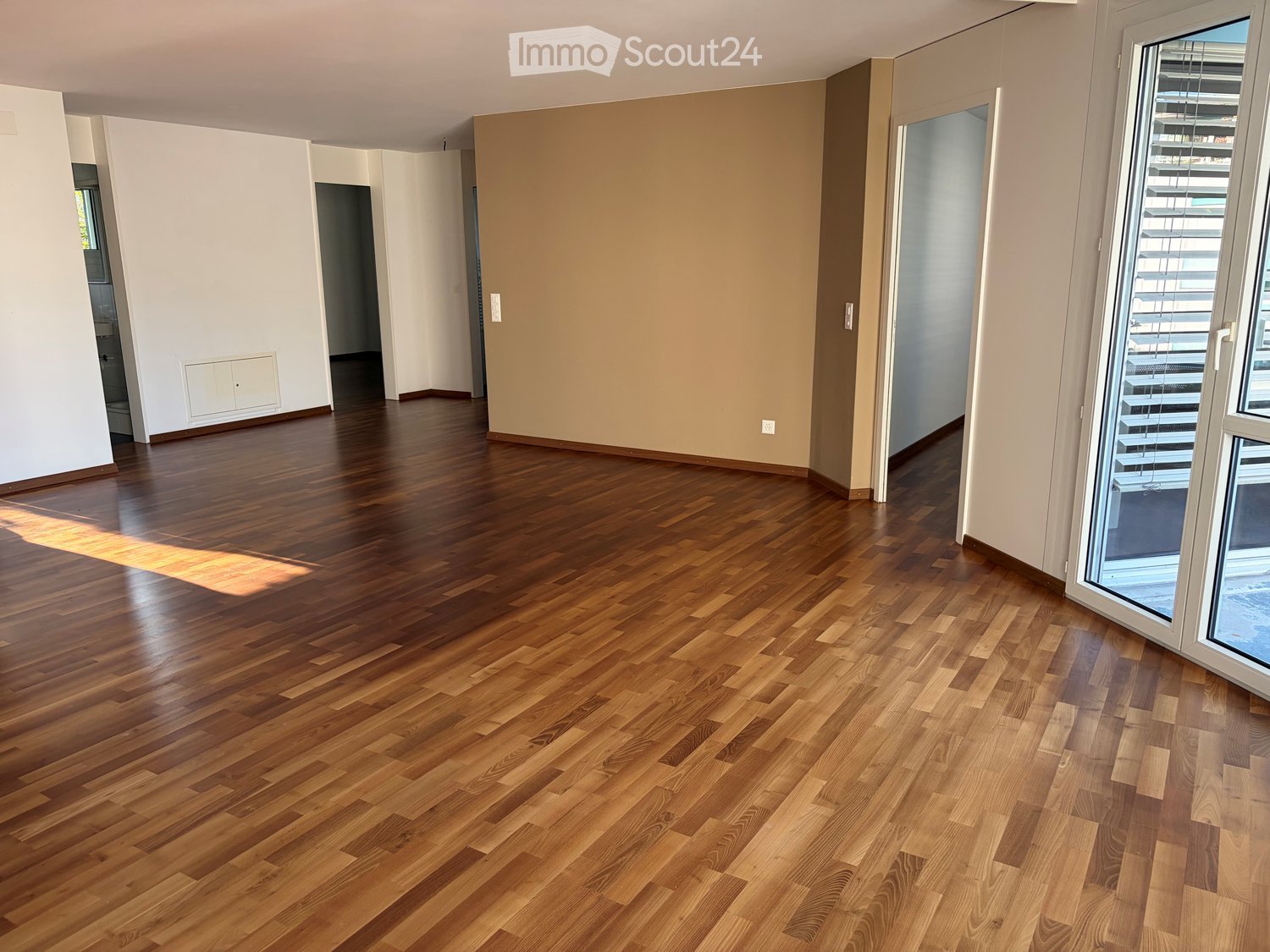 Empty room with polished wooden flooring, white walls, and large windows with blinds. No furniture or equipment present.