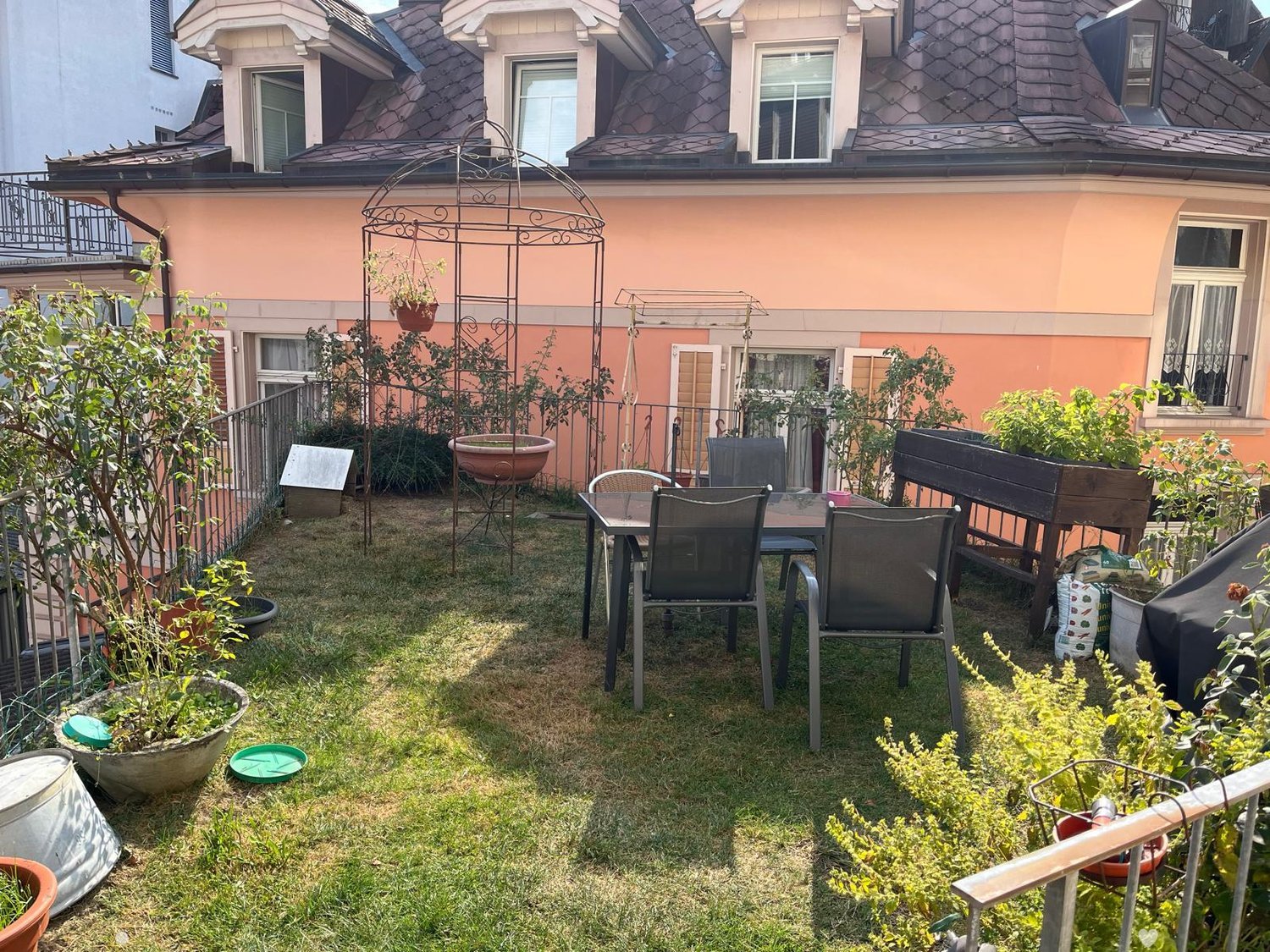Small garden area with a table and chairs, surrounded by potted plants and a wooden bench. The space is enclosed by a metal fence and features a metal garden trellis.