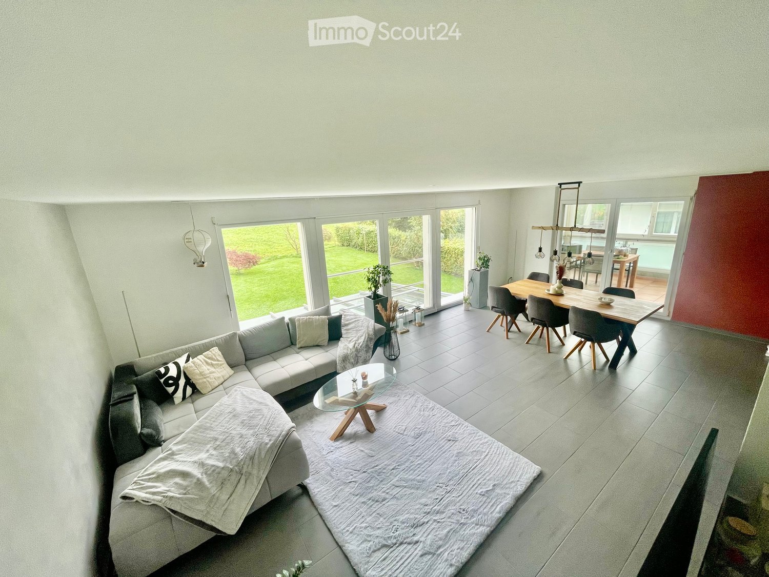 Modern living and dining area with large windows, grey sectional sofa, glass coffee table, dining table with chairs, and decorative vases.