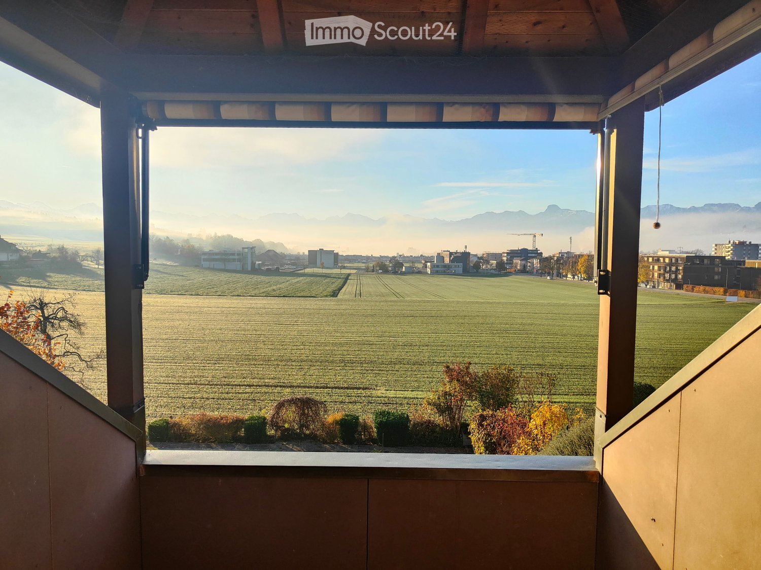 Balcony with wooden railings and a view of a rural landscape, fields, and distant mountains.