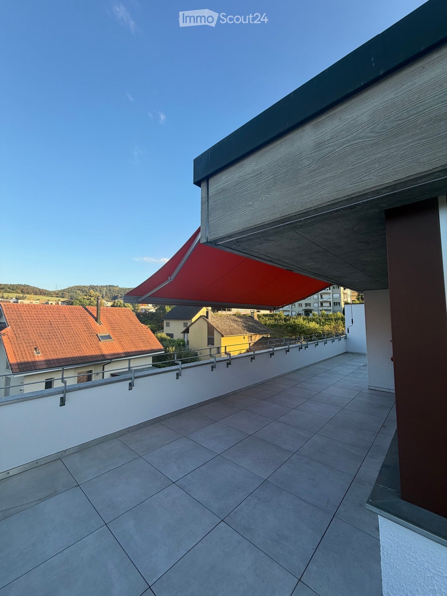 Balcony with tiled floor, white walls, metal railing, and a red retractable awning extending from the roof.