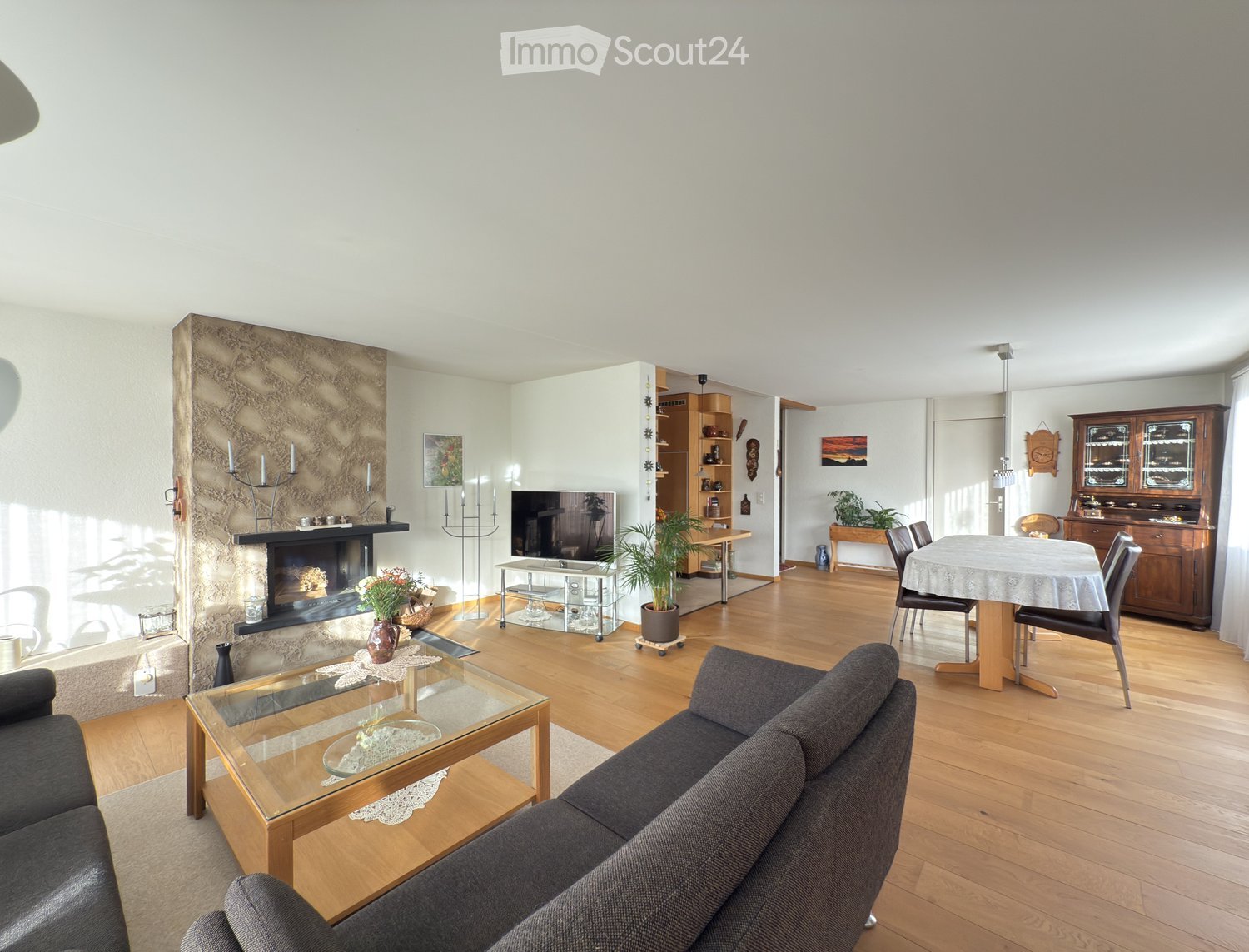 Bright living room with wooden flooring, a dark grey sofa, a glass coffee table, a fireplace with candles, a TV stand, a dining area with a table and chairs, and a wooden cabinet.