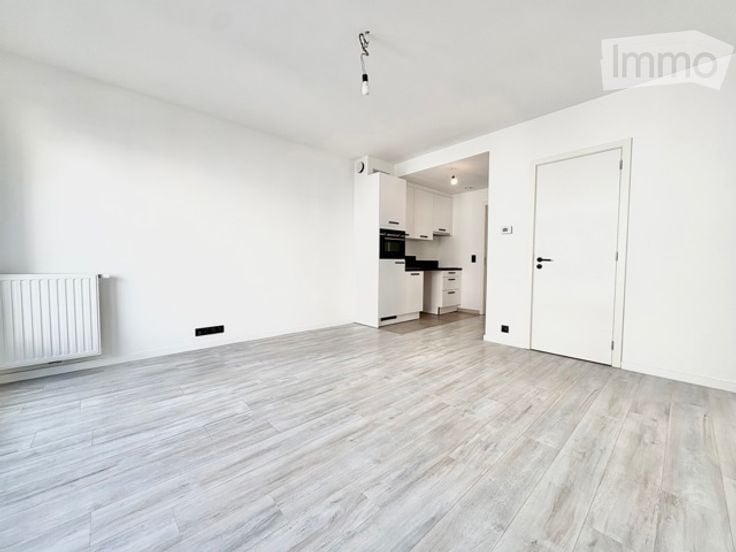 Empty room with white walls, light wooden flooring, and a radiator on one wall. A doorway leads to a small kitchen area with white cabinets and a black oven.