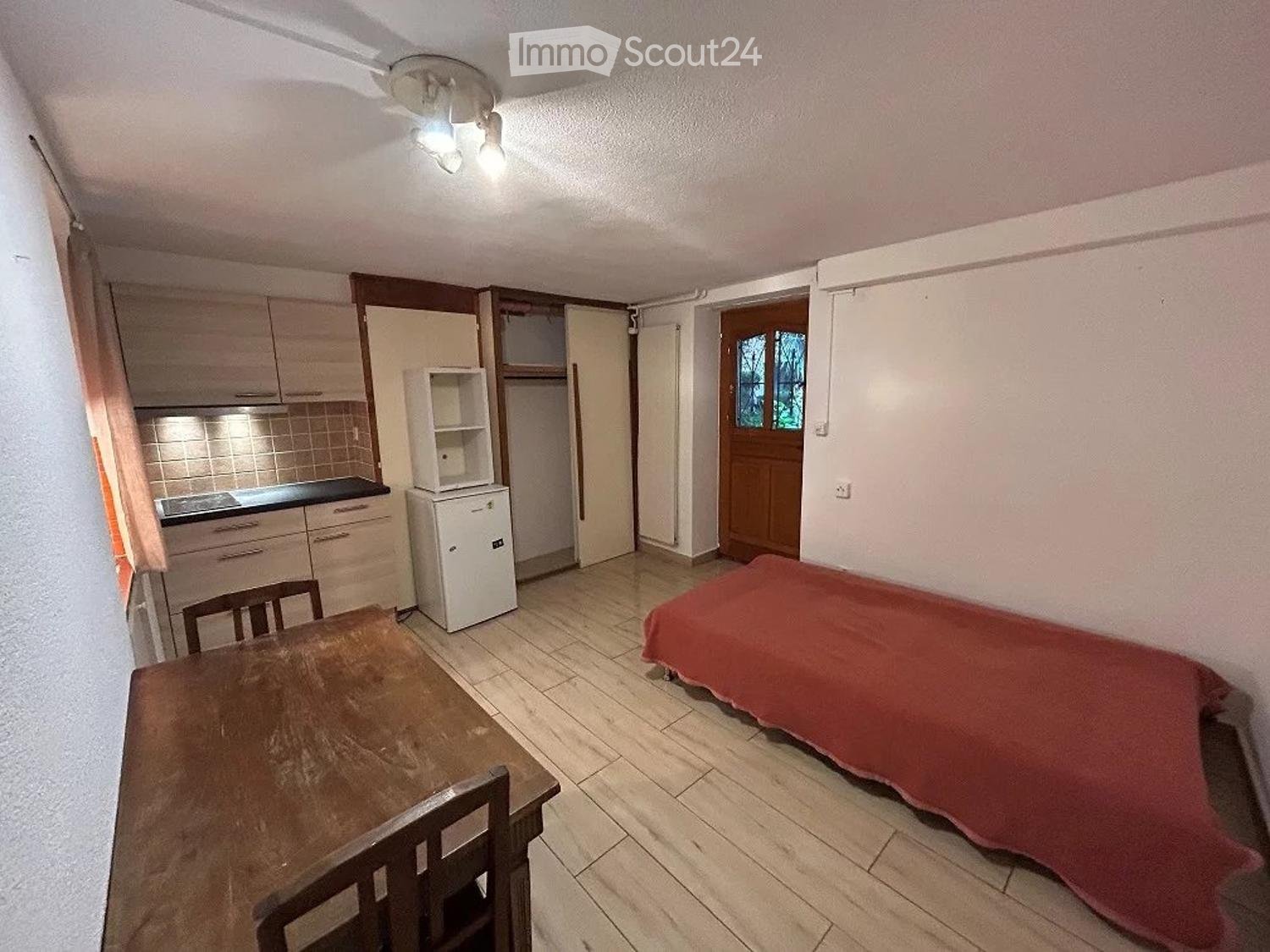 A compact studio apartment with a bed, a small table and chairs, a refrigerator, and a kitchenette area with cabinets and a countertop.