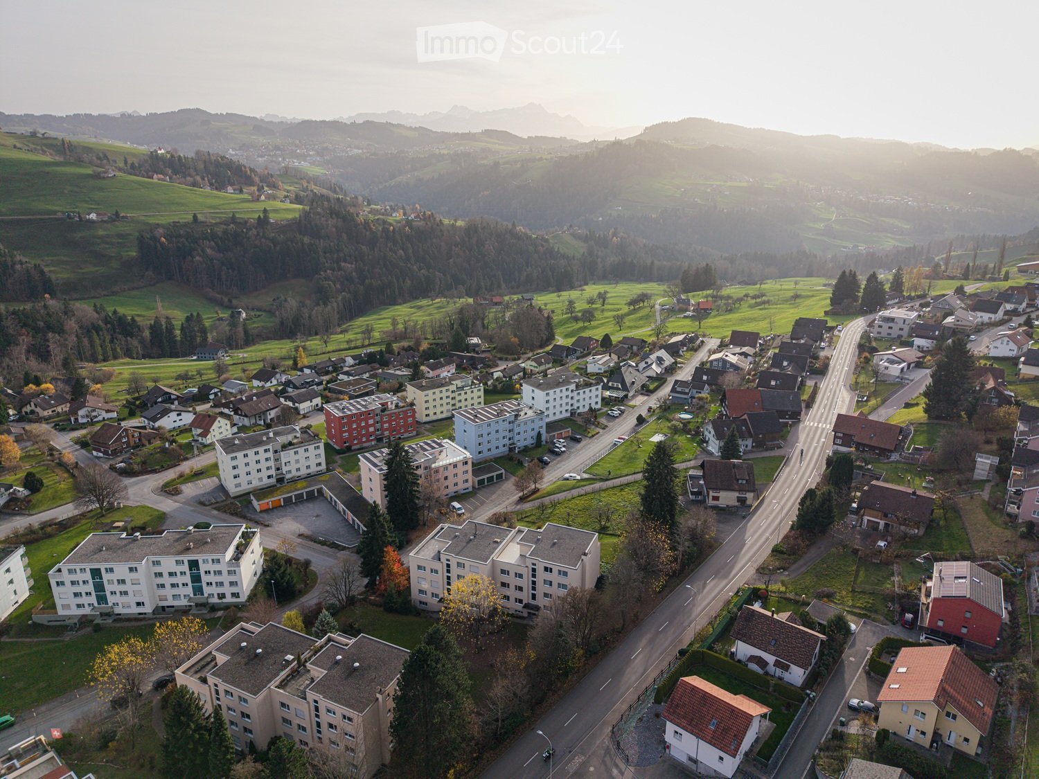 Aerial view of a suburban neighborhood with residential buildings, roads, and surrounding green fields and hills.