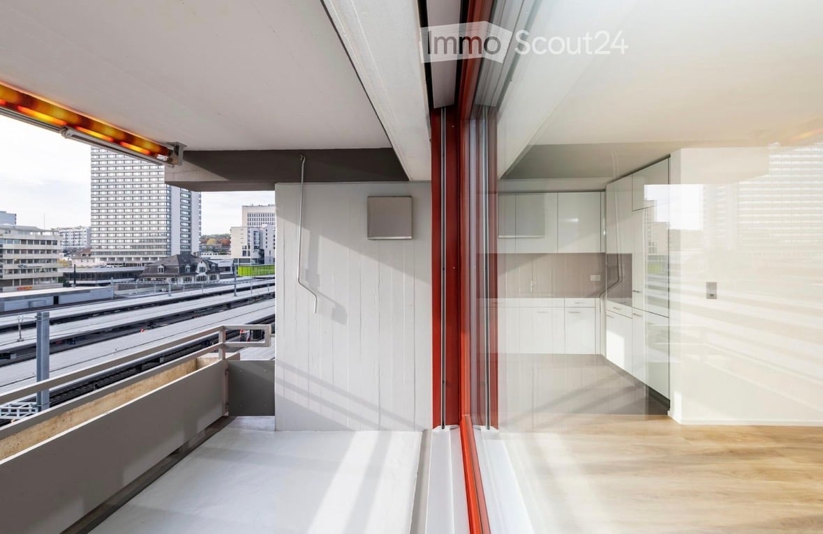 Modern balcony with large glass windows, wooden flooring, and a view of the cityscape and train tracks.