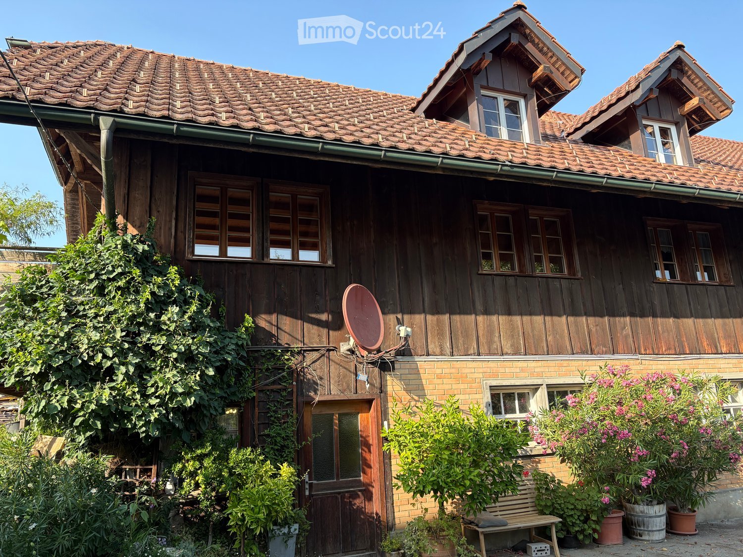 Two-story wooden house with tiled roof, multiple windows, and a satellite dish on the side. The house is surrounded by greenery and potted plants, with a wooden bench near the entrance.