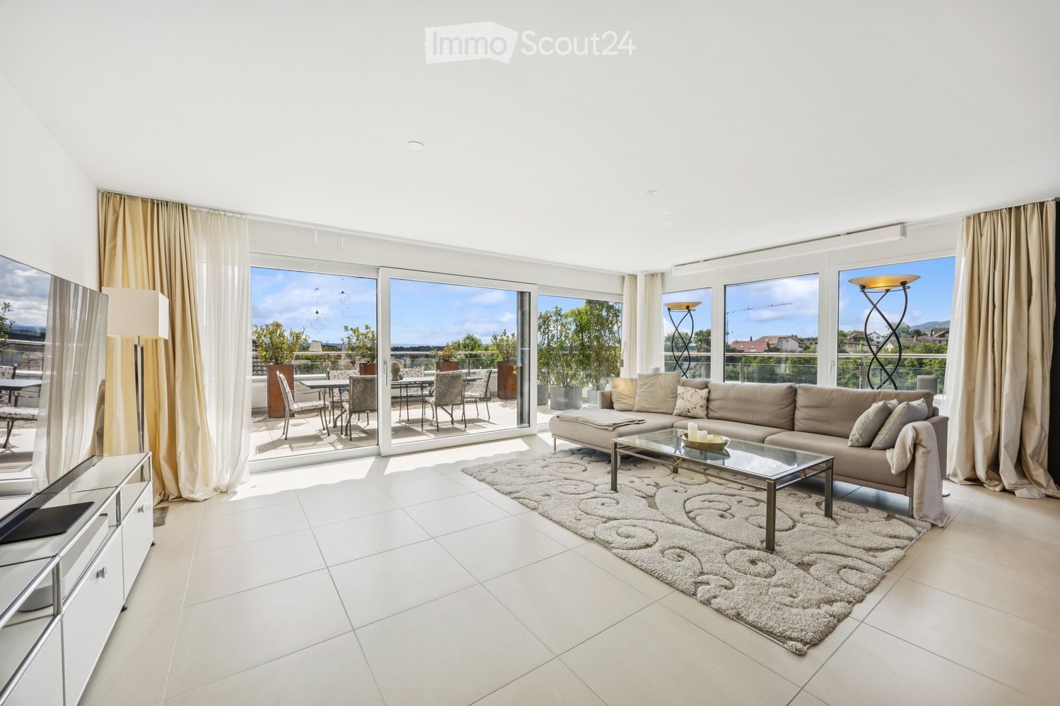 Modern living room with large windows and a balcony view, featuring a beige sofa, glass coffee table, floor lamp, TV stand, and dining area with chairs and table.