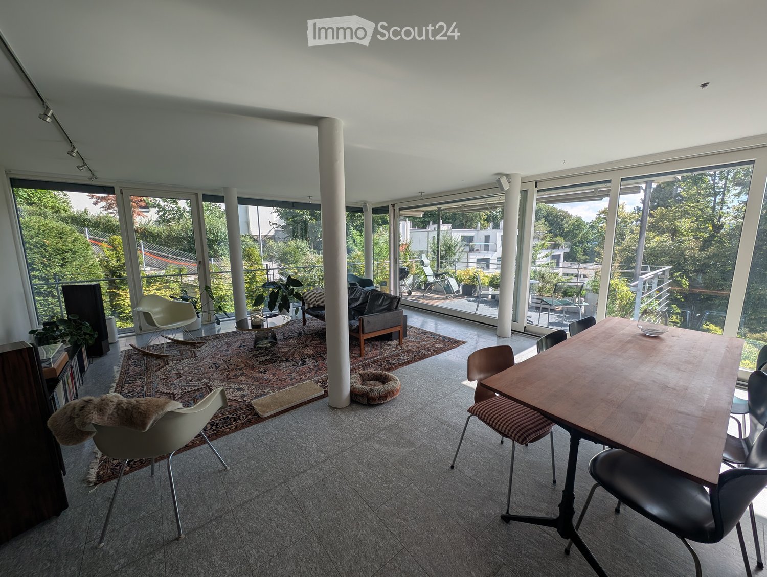 Bright living and dining area with large windows and a balcony, featuring a sofa, dining table, chairs, and plants.