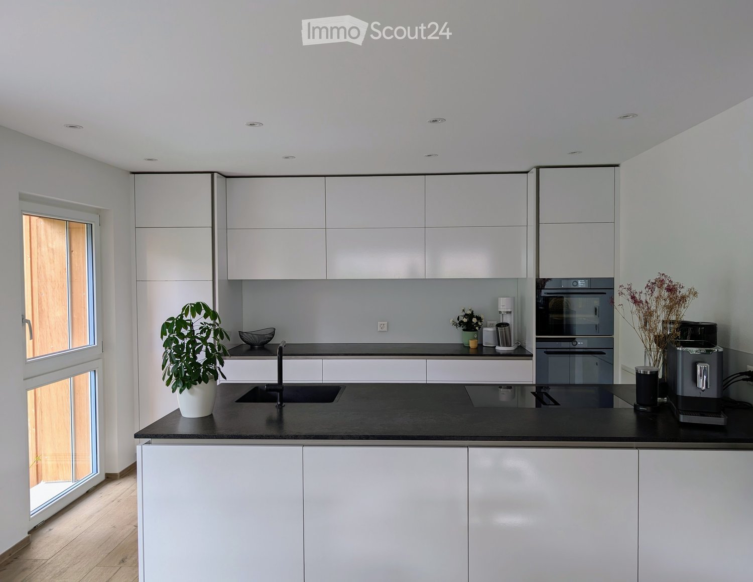 Modern kitchen with white cabinets, black countertop, stainless steel appliances, and a large window. Includes a sink, faucet, oven, coffee machine, and decorative items.