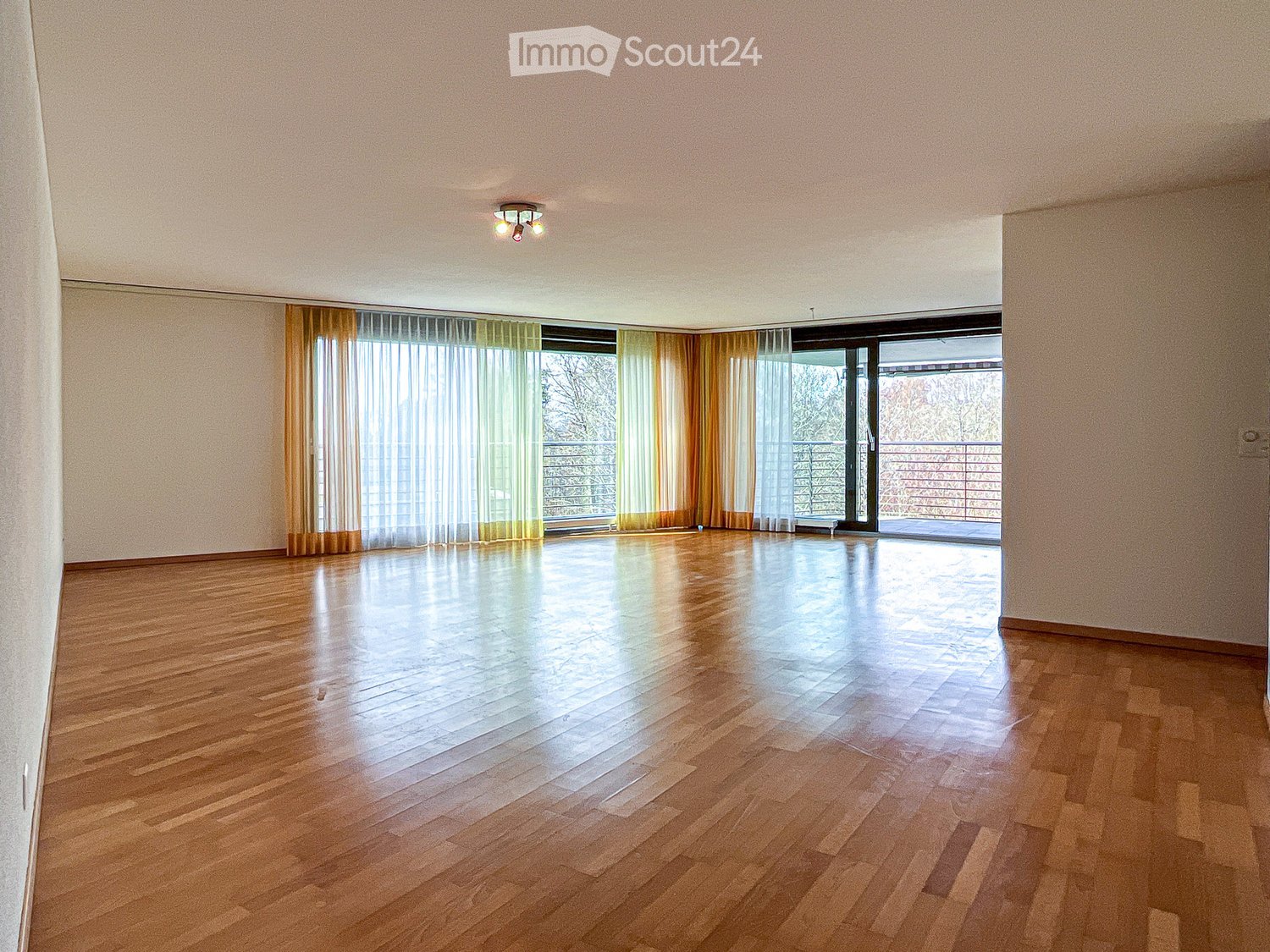 Empty room with wooden flooring, large windows with curtains, and a sliding glass door leading to a balcony.