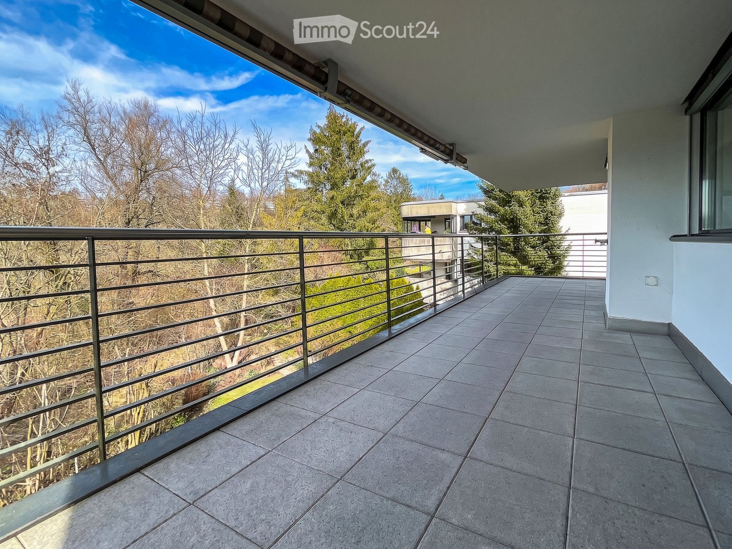 Balcony with tiled floor, metal railing, and a view of trees and a building in the background.