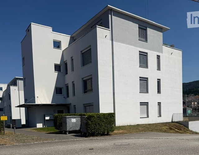 Modern, multi-story white building with rectangular windows and a flat roof, surrounded by a small landscaped area with hedges and a trash bin.