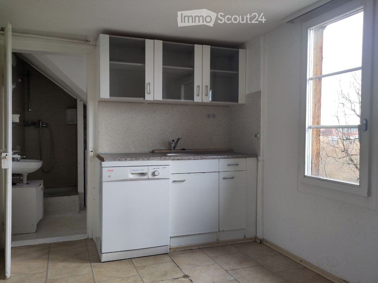 A small, modern kitchen with white cabinets, a dishwasher, a sink, and a mirror above the sink. The room has tiled flooring and a window letting in natural light.