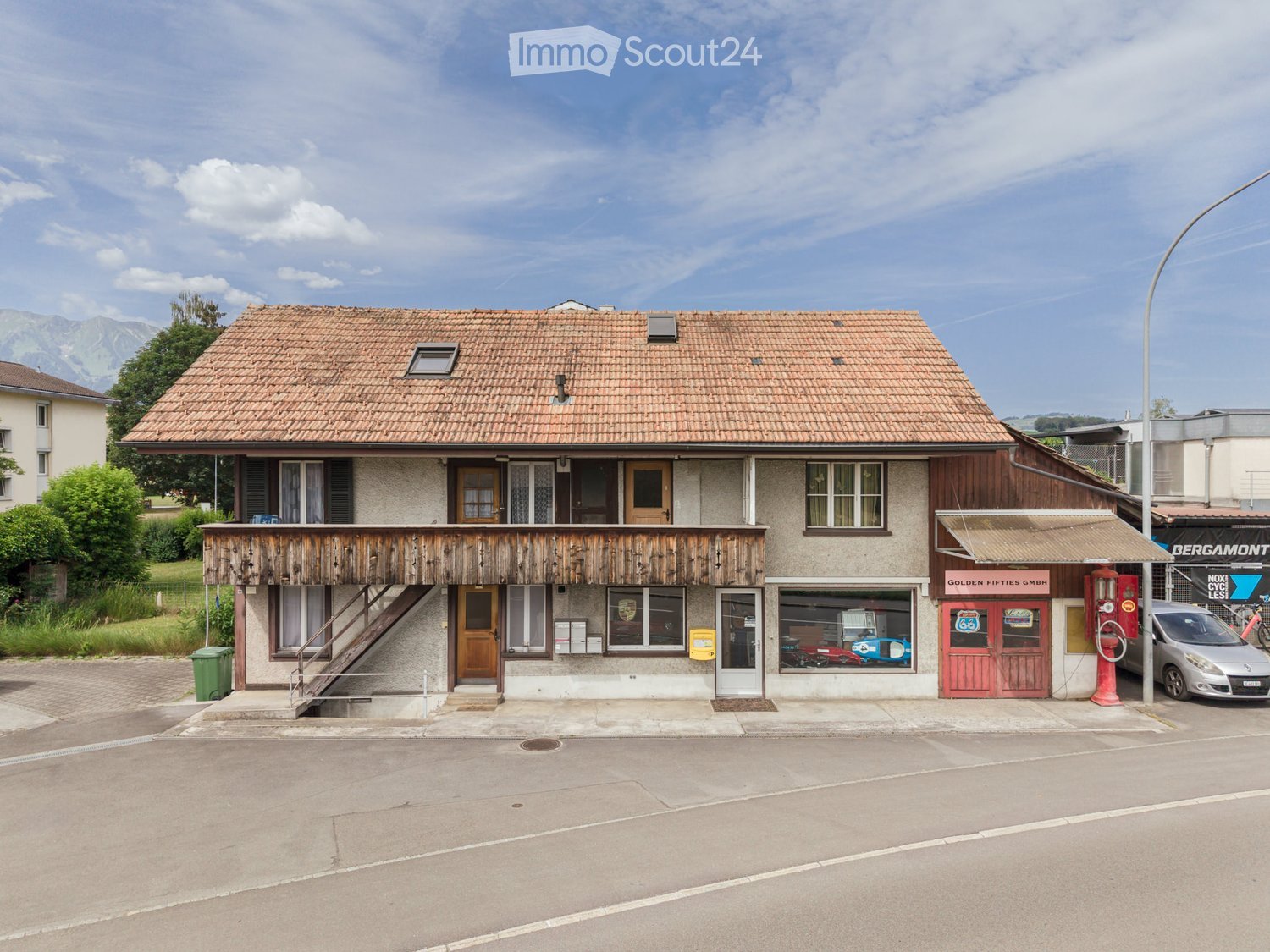Two-story building with a tiled roof, wooden balcony, and storefronts including a car display and a gas pump.