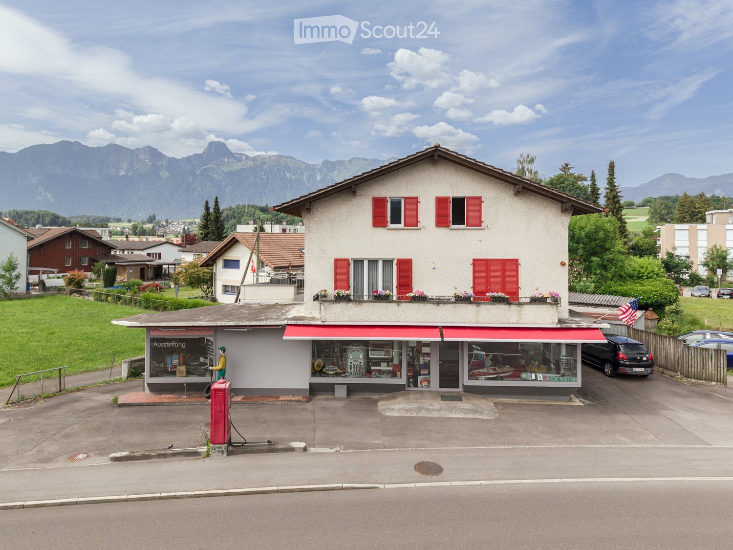 Two-story house with red shutters, a balcony with flower boxes, and a storefront with a red awning. The house is situated in a residential area with mountains in the background.