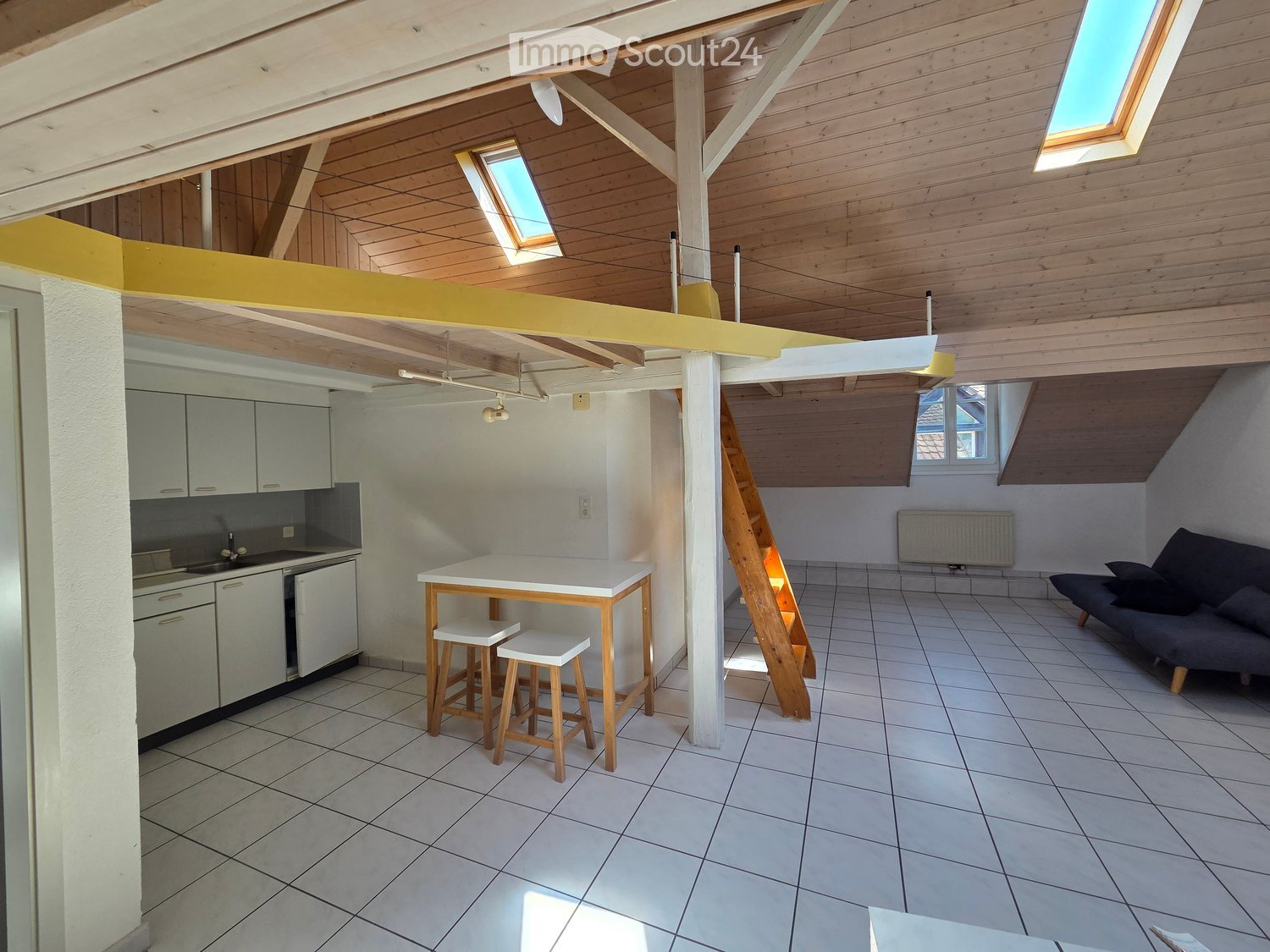 Open-plan living and kitchen area with white tiled flooring, wooden ceiling beams, skylights, a grey sofa, a white dining table with two stools, and a fitted kitchen with cabinets and a sink.