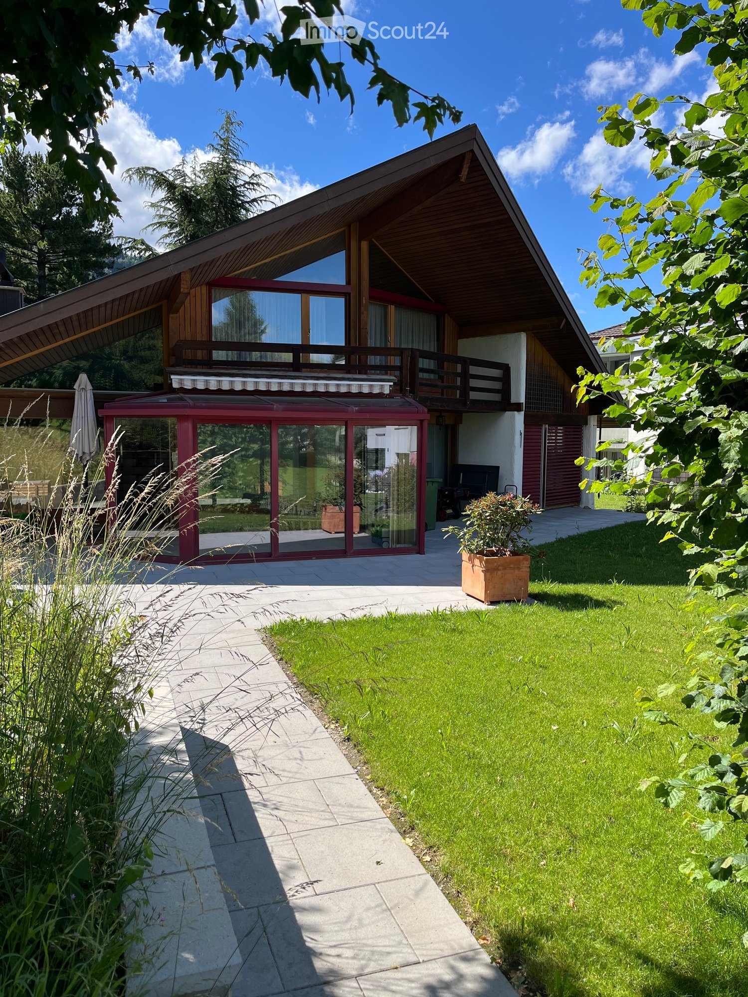 Modern two-story house with a sloped wooden roof, large windows, and a balcony. The house features a patio area with glass walls, a small garden, and a well-maintained lawn.