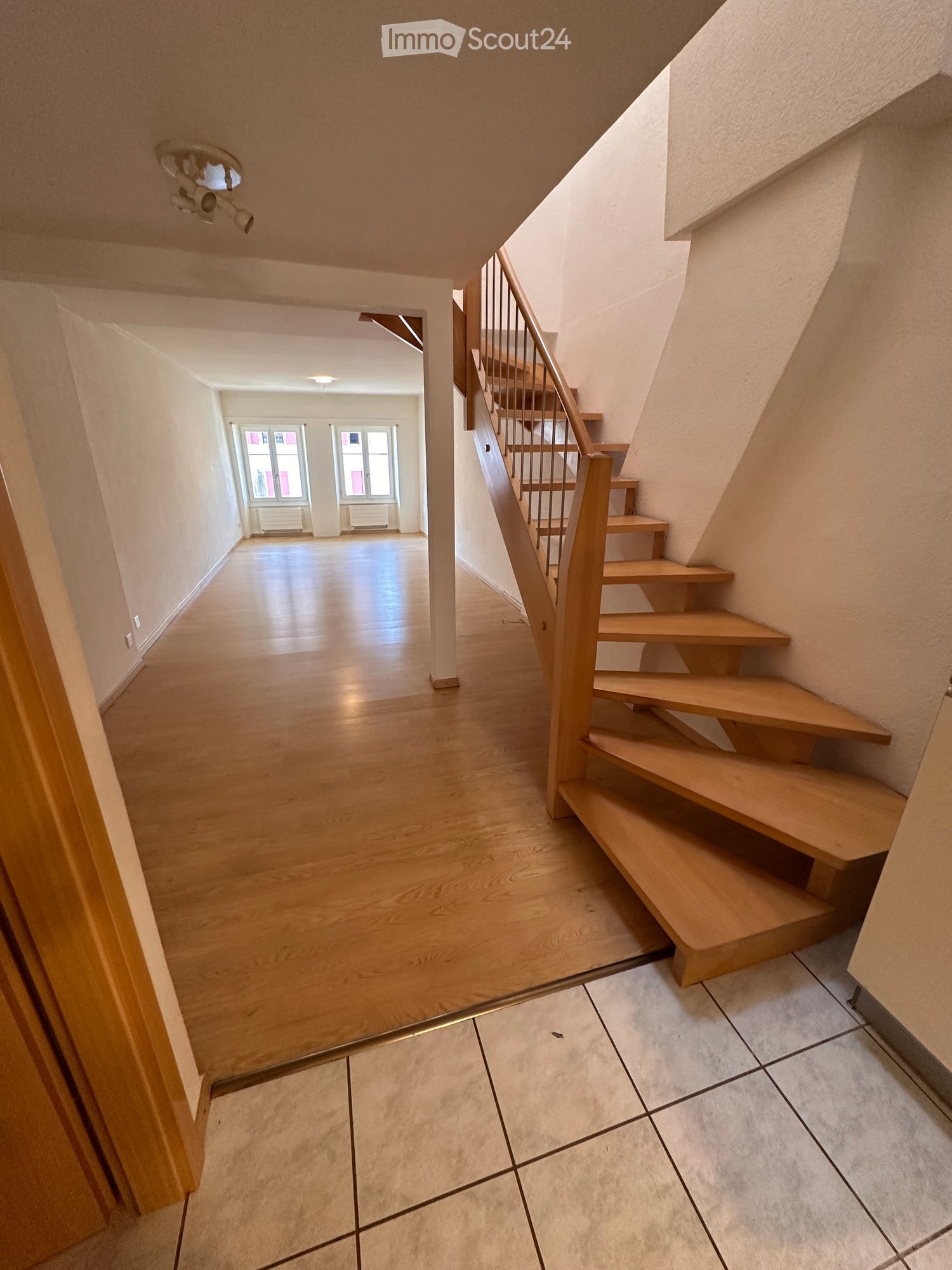Spacious hallway with wooden flooring, a staircase with wooden steps and a metal railing, and a view into an adjacent room with large windows and a radiator.