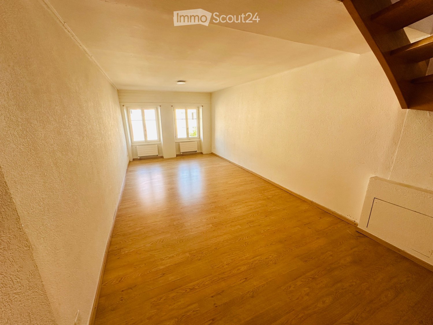 Empty hallway with wooden flooring, white walls, and two windows at the far end.