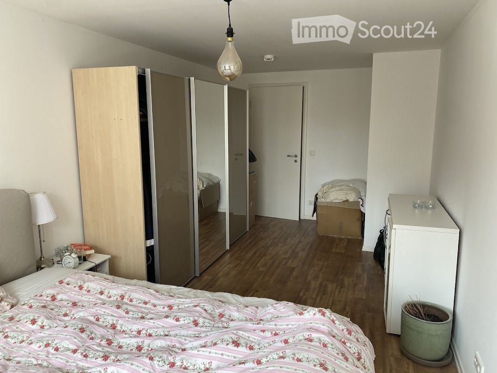 Cozy bedroom with wooden flooring, a neatly made bed with floral bedding, a nightstand with a lamp, a wardrobe, and a small refrigerator. A potted plant is placed near the refrigerator.