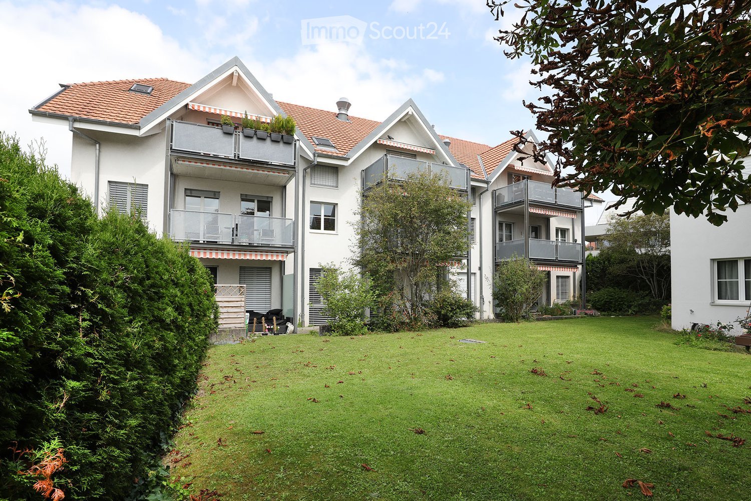 A modern two-story house with white exterior, red-tiled roof, multiple balconies, and a well-maintained lawn in front.