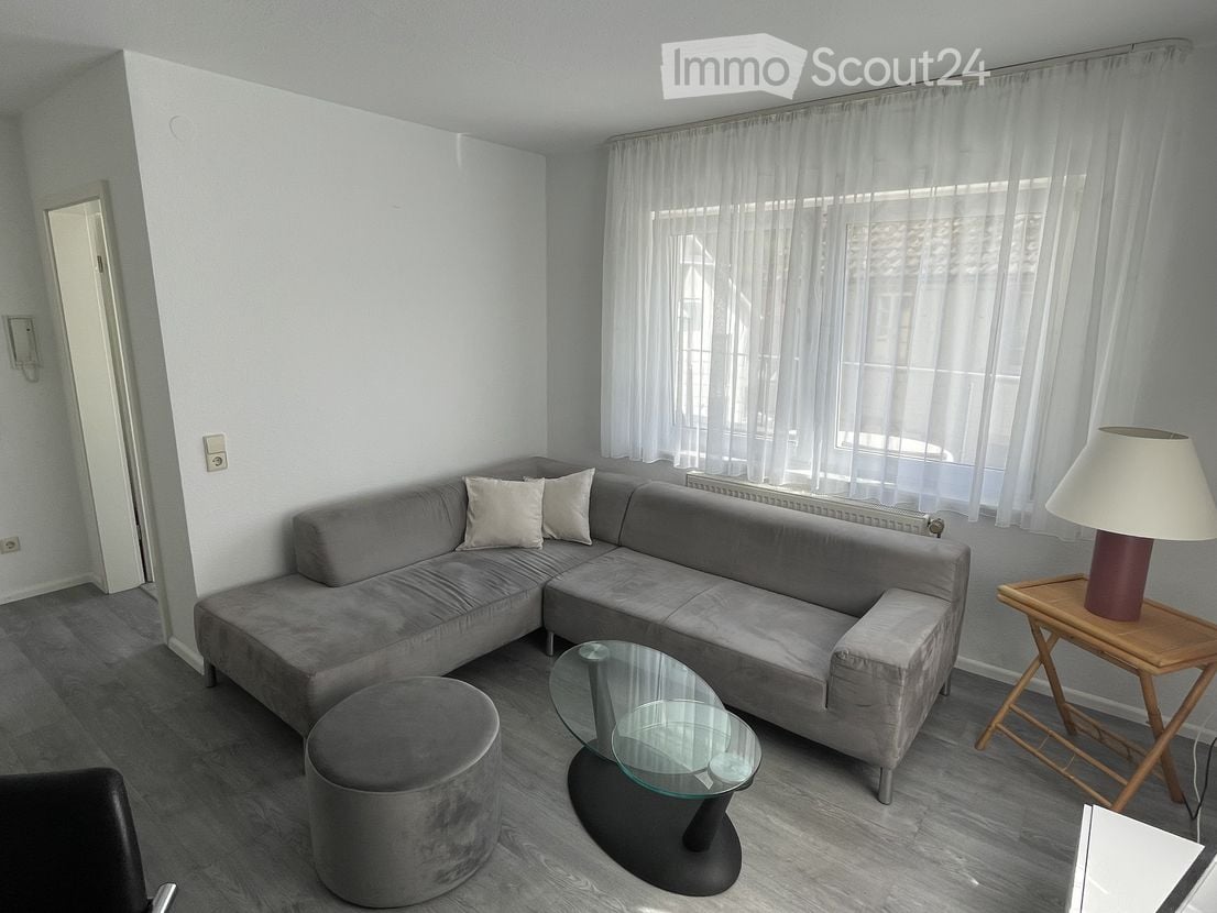 Modern living room with grey sectional sofa, ottoman, glass coffee table, side table with lamp, and large windows with white curtains.