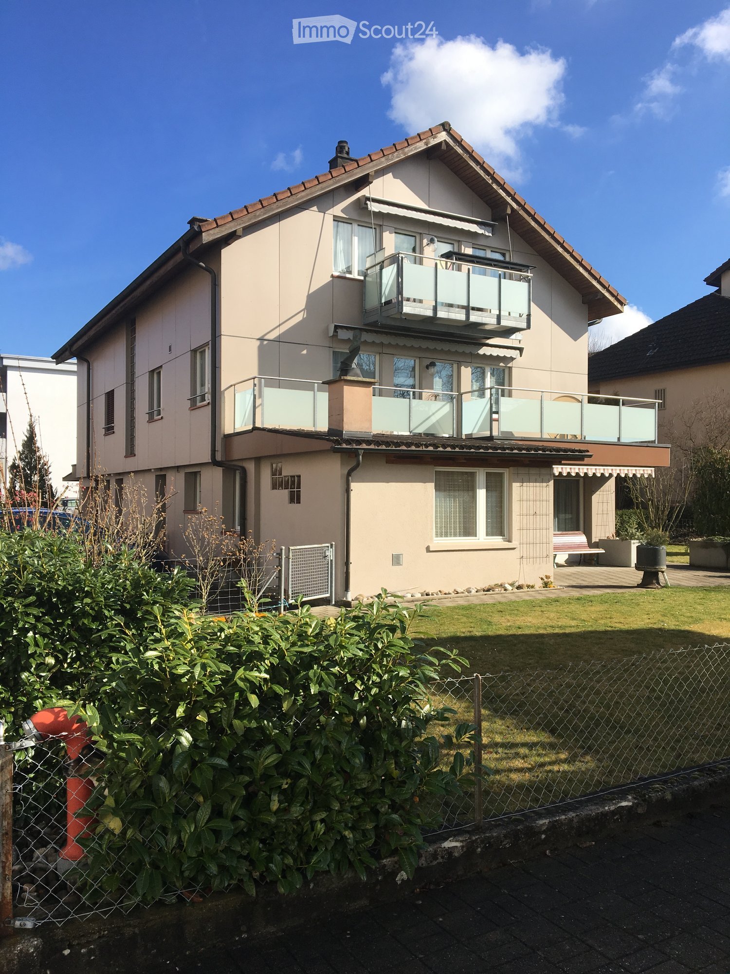 2-story residential building with beige exterior, red tile roof, balconies, and a fenced yard with vegetation.