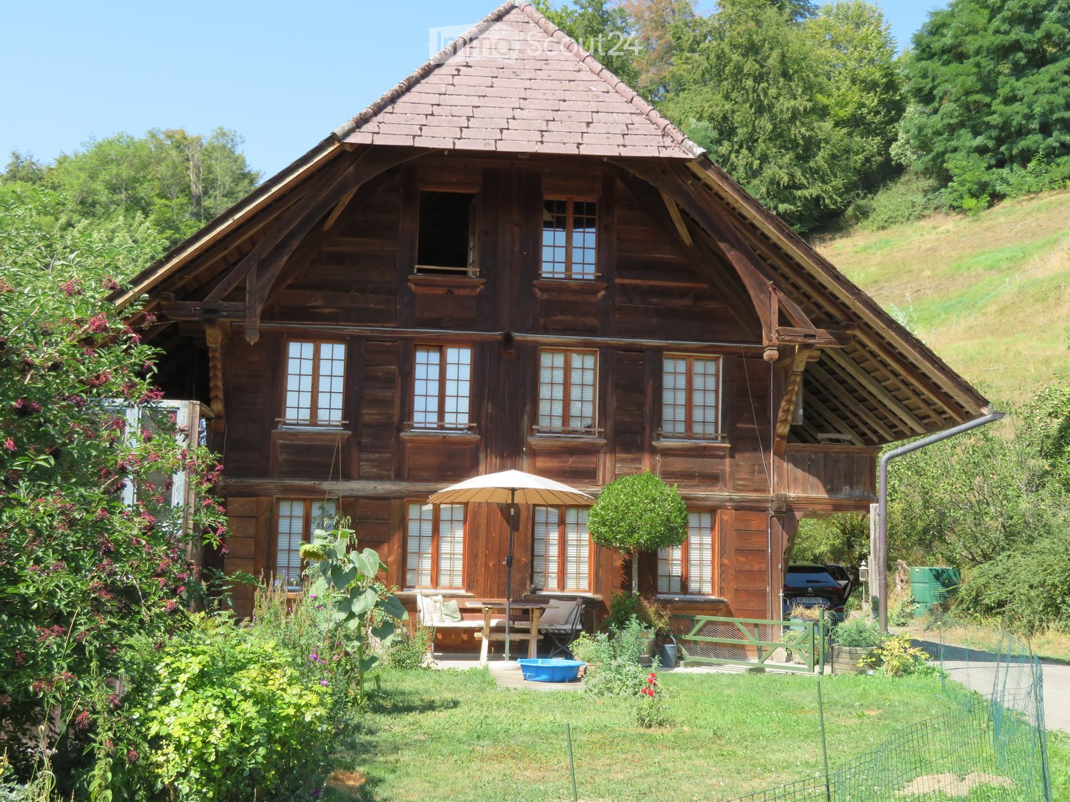 Traditional wooden house with a steep, triangular roof and multiple windows, surrounded by a well-maintained garden with outdoor seating and a parked car.