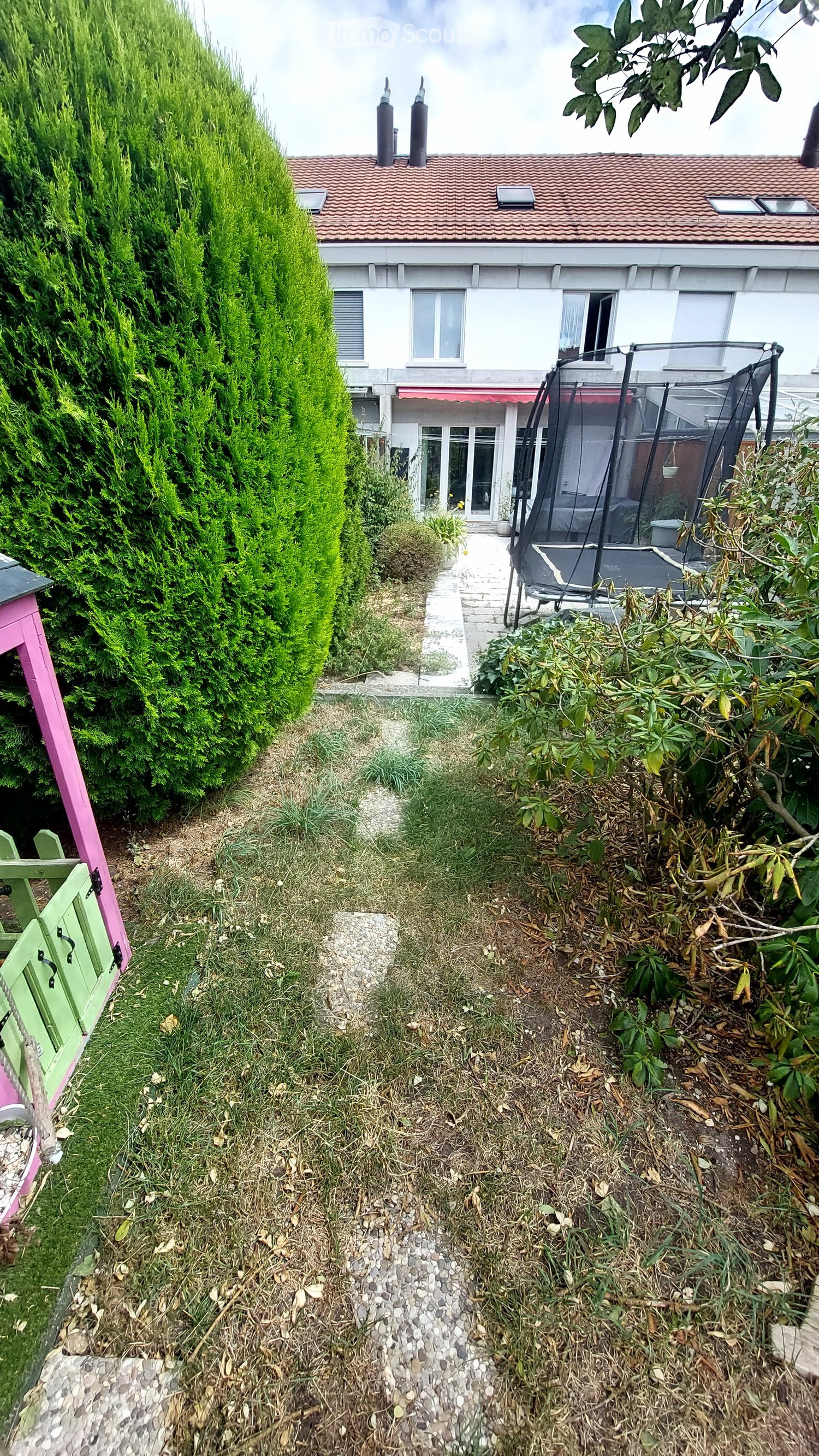 A narrow garden path leads to a house with a trampoline in the background. The garden features a tall, trimmed hedge on one side and various plants and shrubs on the other.
