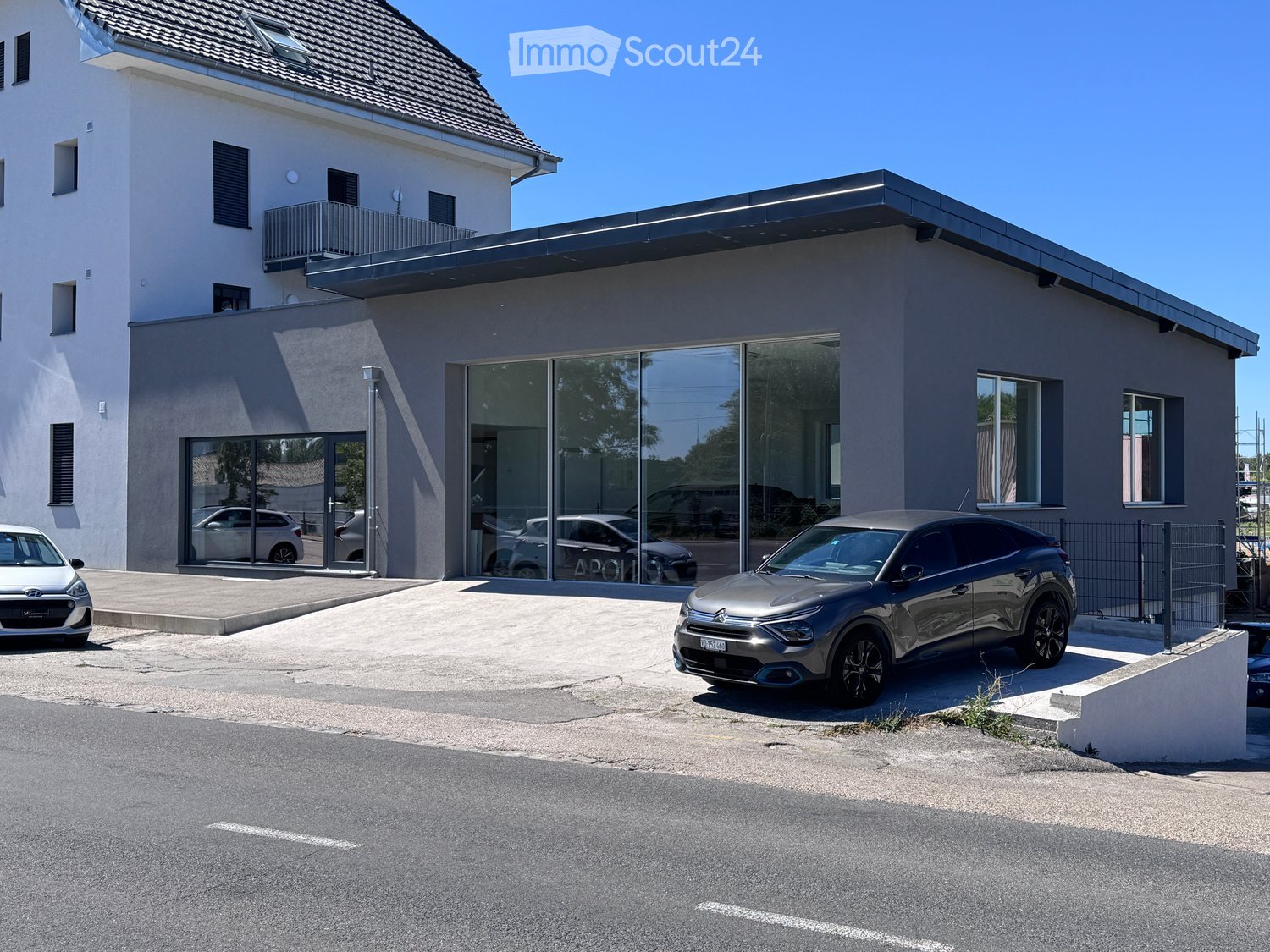 Modern two-story building with a balcony and large glass windows, with several cars parked in front.