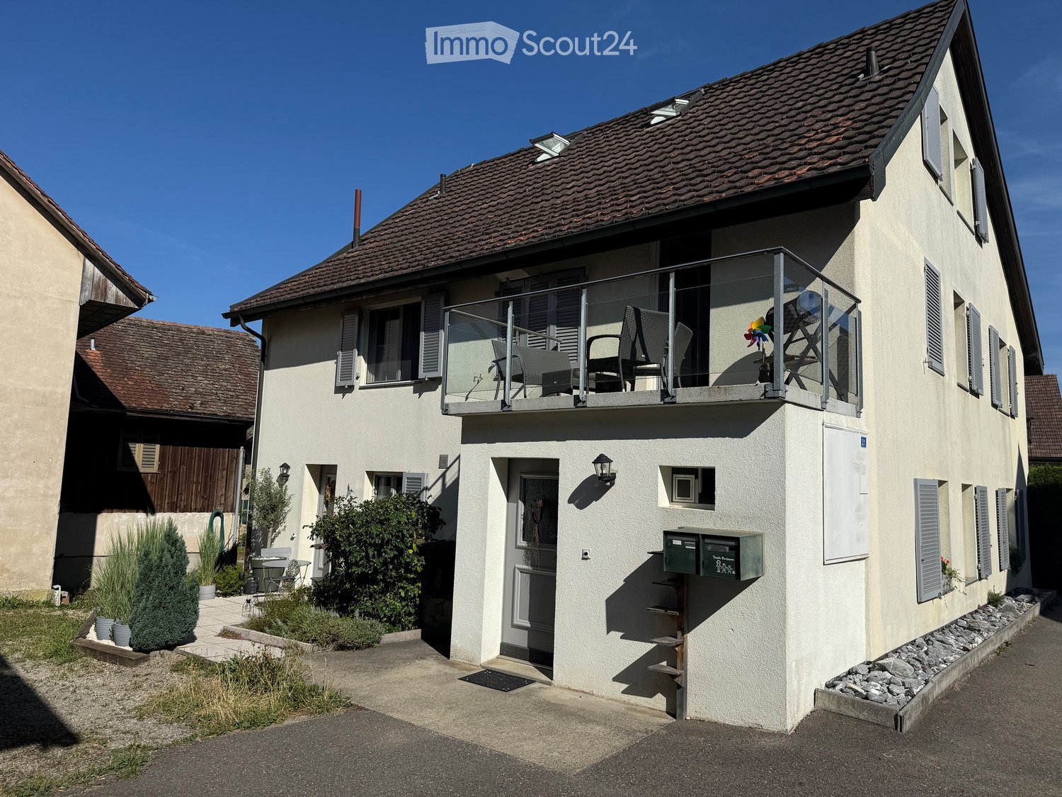 Two-story house with beige exterior, tiled roof, balcony with chairs, small garden, and mailbox.
