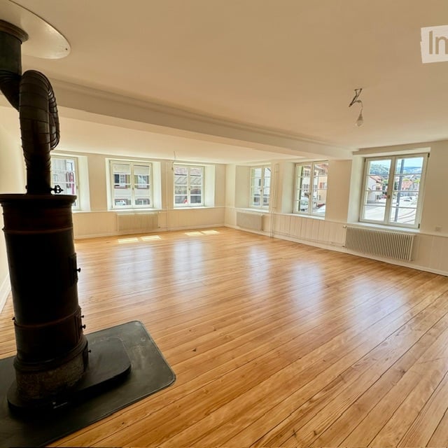 Empty room with wooden flooring, large windows, and a central heating stove. No furniture or equipment present.