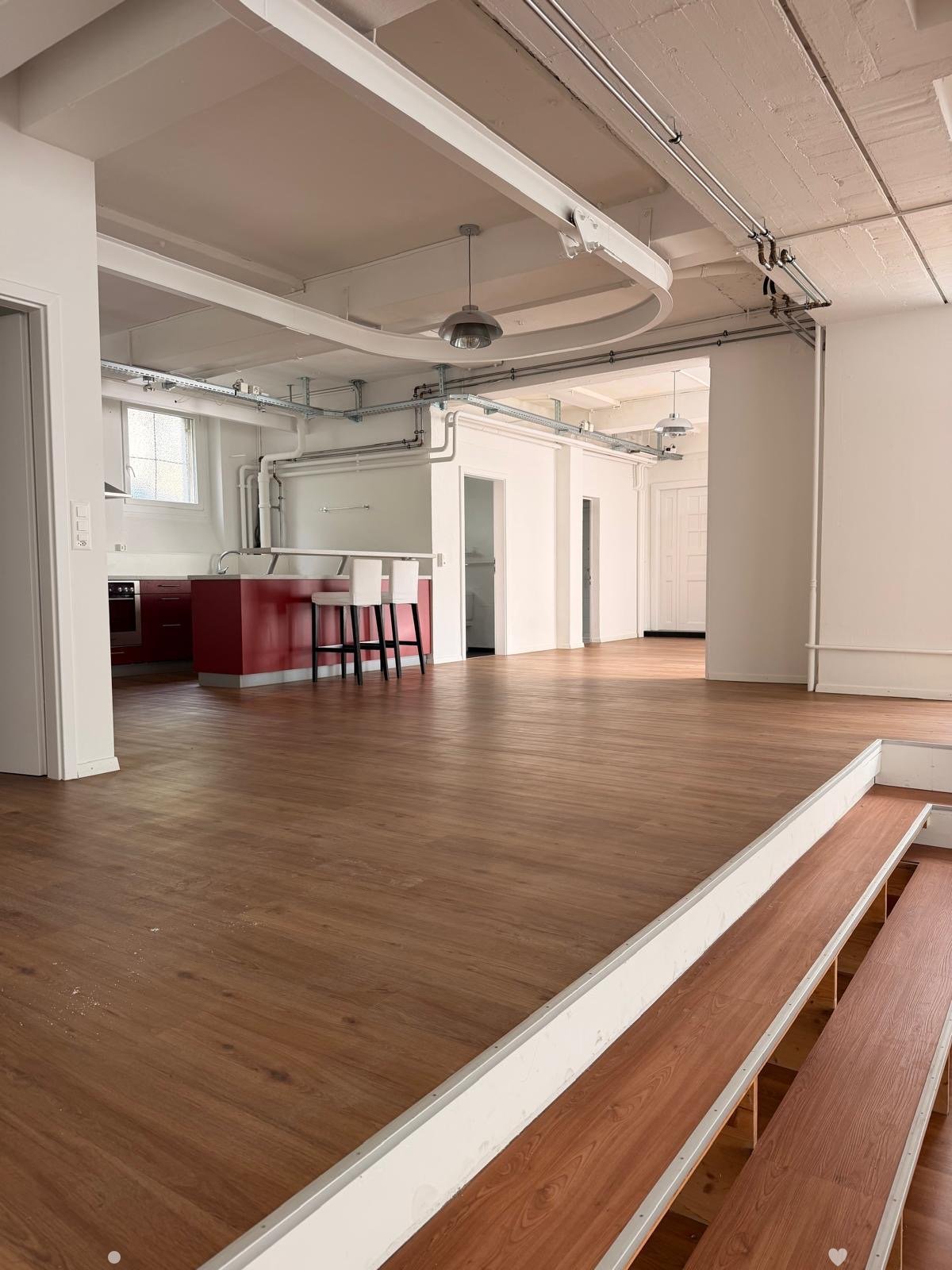 Open-plan space with wooden flooring, exposed pipes and ductwork, a small kitchen area with a red island, two stools, and an oven.