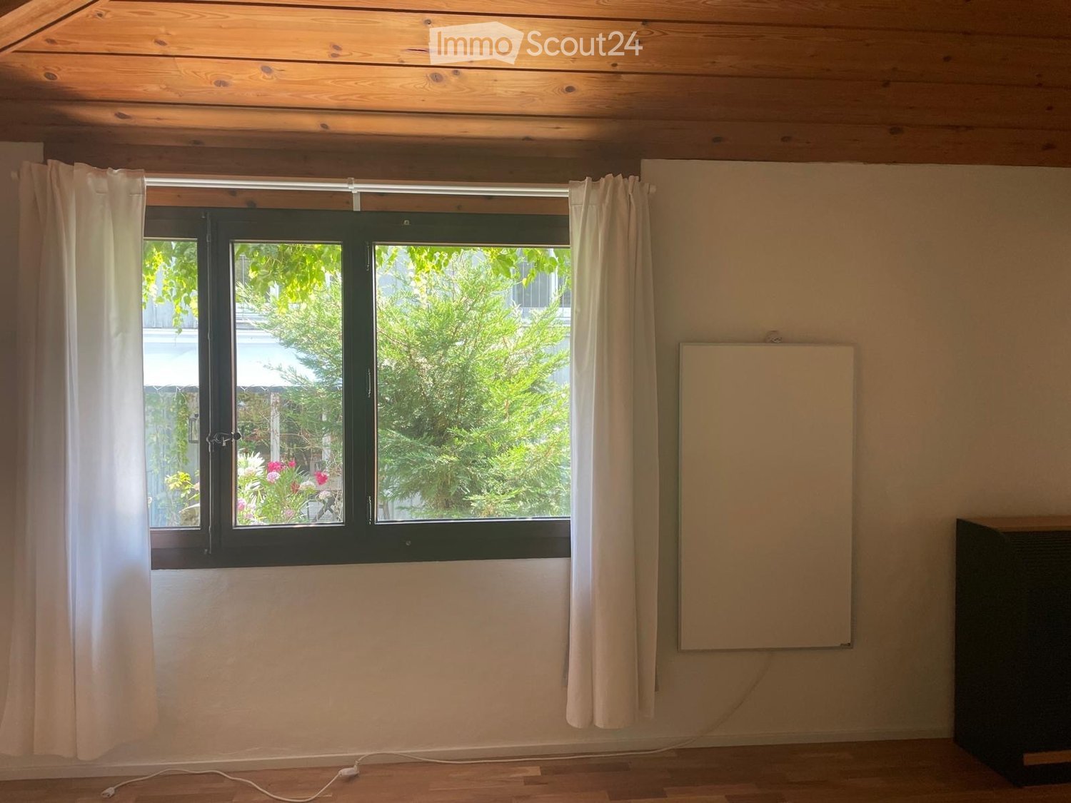 Empty room with wooden floor, large window with white curtains, and a whiteboard on the wall.