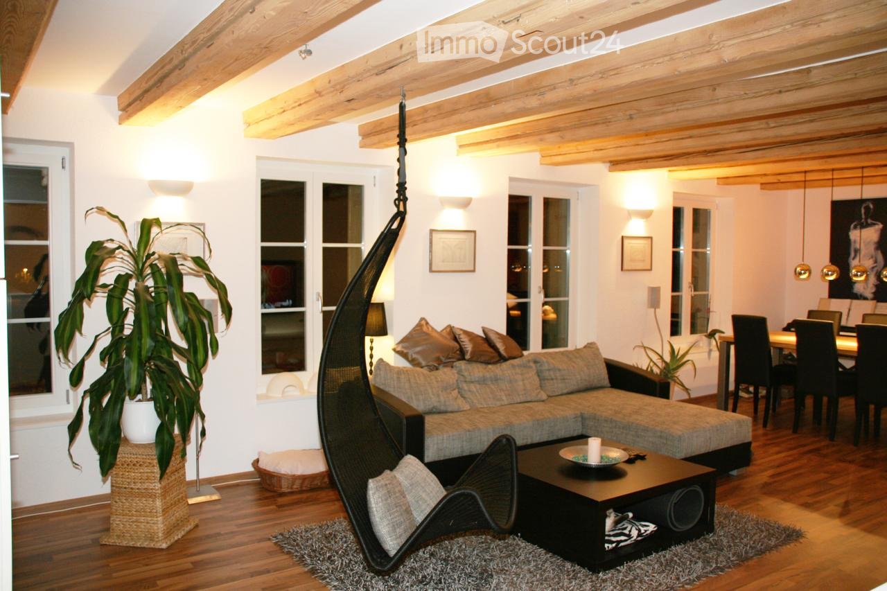 Living room with exposed wooden beams, large windows, a grey sectional sofa, a black wicker hanging chair, a coffee table, and a dining area with chairs and a table.