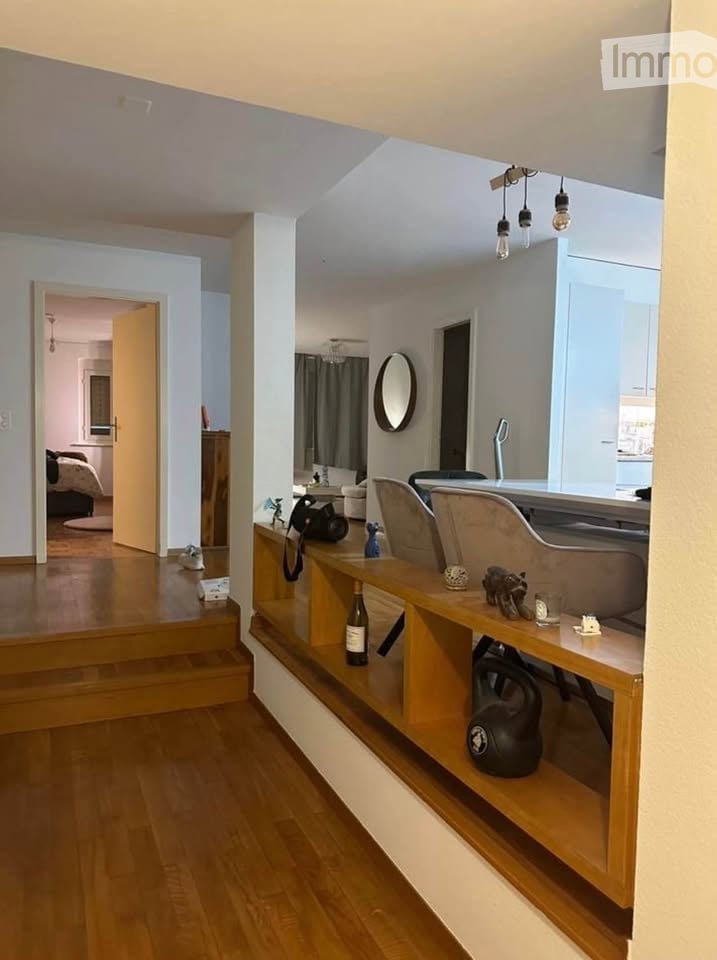 Modern open-plan living and dining area with wooden flooring, built-in shelving, a sofa, chairs, a dining table, and decorative items including a mirror and a bottle on the shelf.