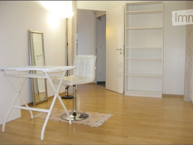 A modern, minimalist room with a white leather chair, a small white table, a mirror, and a white shelf unit. The floor is light wood, and the walls are white.
