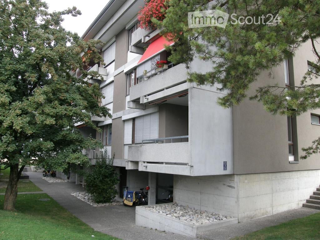 Modern multi-story apartment building with balconies, greenery, and a small parking area.