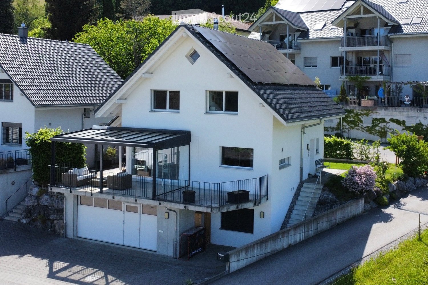 A modern two-story house with white exterior walls, grey tiled roof, large windows, and a covered patio area with seating. The house features a garage and is surrounded by greenery and neighboring houses.
