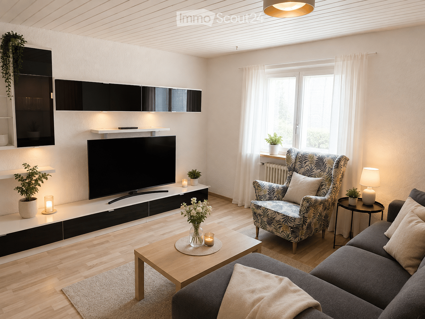 Modern living room with a grey sectional sofa, patterned armchair, wooden coffee table, flat-screen TV on a black and white entertainment unit, side table with lamp, and decorative plants.
