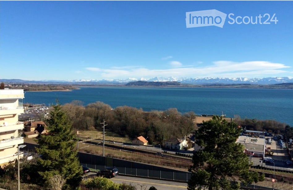 Panoramic view of a lake with snow-capped mountains in the background, surrounded by trees and buildings.