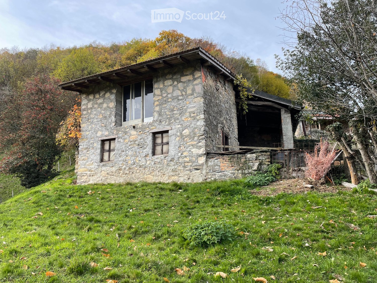 Stone house with a slanted roof, multiple windows, and a small porch area surrounded by greenery and trees.