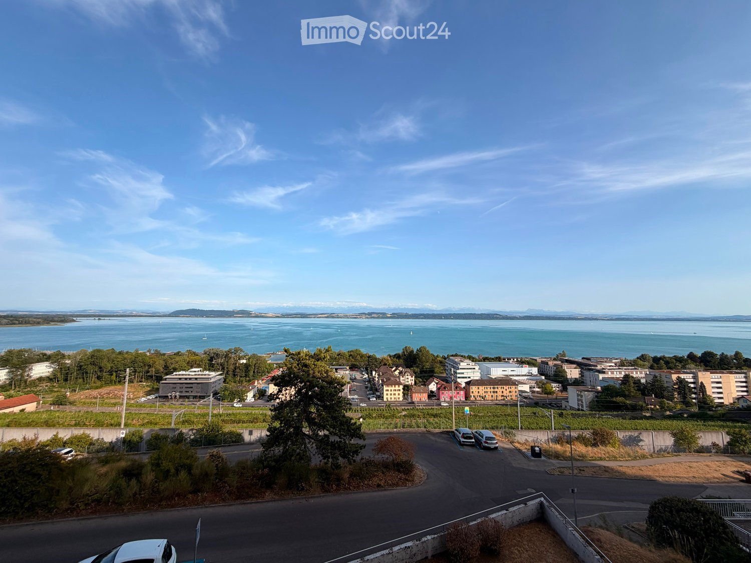 A panoramic view of a coastal city with a body of water, buildings, and a parking area in the foreground.