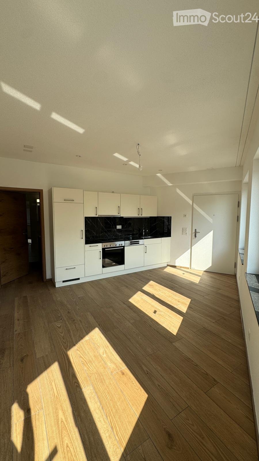 Modern kitchen with white cabinets, a built-in oven, and light wooden flooring. Sunlight streams in, casting shadows on the floor.